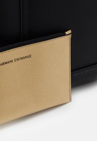 Pochette en cuir beige avec fermeture éclair noire partiellement à l'intérieur d'un sac en cuir texturé noir, les deux portant le logo "Armani Exchange".