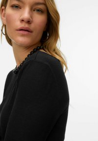 Zwarte top met lange mouwen en een ronde hals, met een gladde textuur en subtiele naaddetails. Het model draagt een grove zwarte kralenhalsketting.