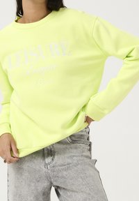 Neon żółta bluza z ściągaczami przy mankietach i dekolcie, z białym napisem "LEISURE League" i rokiem założenia. Noszona z szarymi dżinsami.