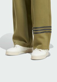  Bílé tenisky spárované s olivově zelenými kalhotami, které mají tři tmavé horizontální pruhy blízko dolních lemů nohavic.