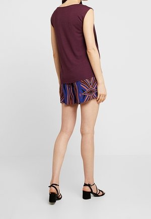 Mujer con una camiseta sin mangas morada oscura, pantalones cortos estampados en azul y naranja, y sandalias negras de tacón ancho con tiras, de pie de espaldas.