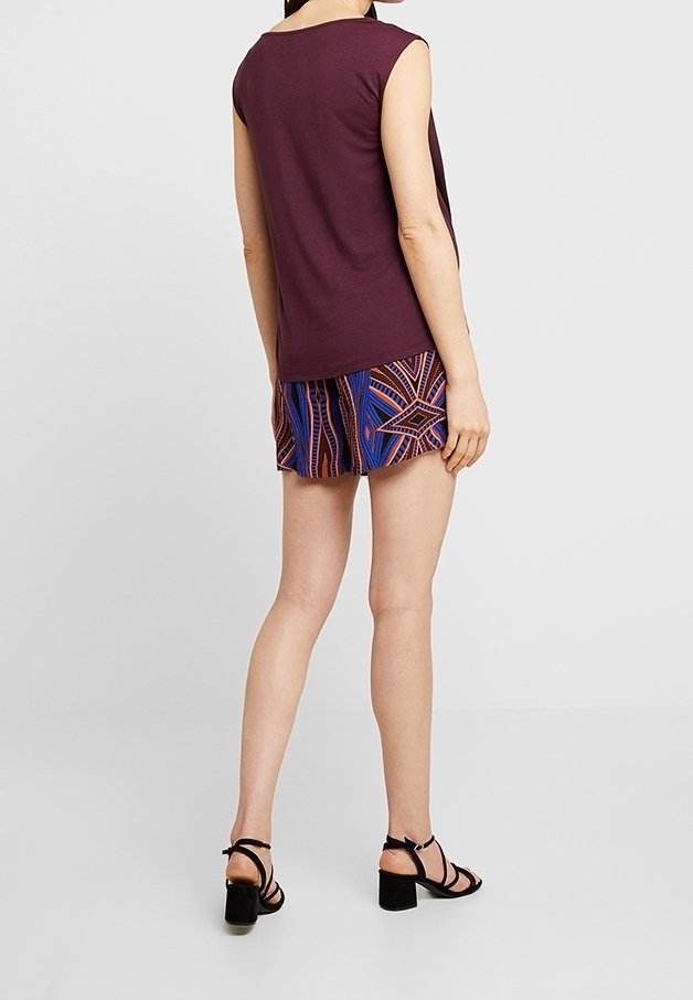 Femme portant un haut sans manches violet foncé, un short à motifs bleu et orange, et des sandales noires à talons blocs avec lanières, debout de dos.