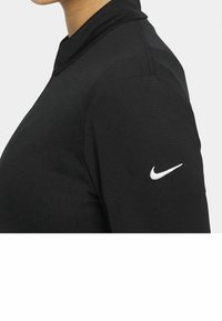 Svart långärmad idrottströja med hög krage bärs av en person, med en vit Nike swoosh-logotyp på den övre ärmen.