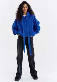 Hanorac electric albastru cu glugă, șnururi lungi și buzunar cangur, asortat cu blugi negri, largi și adidași negri.