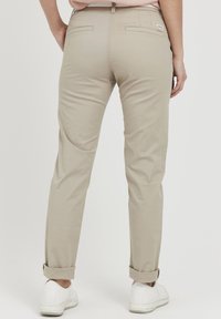 Beige Chinos mit schmaler Passform, aufgerollten Säumen und dezenten Gesäßtaschen. Aus Baumwolle mit glatter Textur. Weiße Sneaker sichtbar.