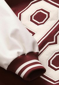 Sweatshirt bordeaux avec une manche en cuir blanc et un grand numéro brodé 98 en crème et bordeaux, avec des poignets côtelés rayés.
