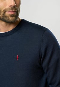 Marineblaues gestricktes Sweatshirt mit einem gerippten Kragen, versehen mit einem kleinen roten Logo auf der Brust und strukturierten Stoffdetails.