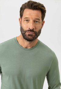 T-shirt vert à manches longues en tissu doux, avec un col rond et une texture lisse, présenté sur un modèle barbu.