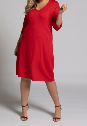 Femme portant une robe rouge mi-longue à col en V avec des manches trois-quarts et des sandales à talons hauts rouges à bride à la cheville, debout sur un sol gris.