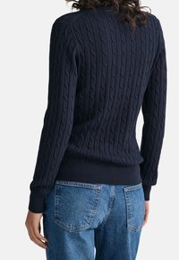 Vrouw in een marineblauwe kabelgebreide trui en blauwe spijkerbroeken, gezien vanaf de achterkant tegen een eenvoudige lichte achtergrond.