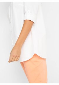 Chemise en lin blanche avec manches retroussées, dotée d'un ourlet incurvé. Associée à un pantalon orange clair, présentant une texture lisse.