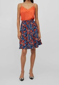 En orange topp med spetskanter, kombinerad med en knälång blå blommig kjol med röda och orange inslag; modell som bär svarta klackar.