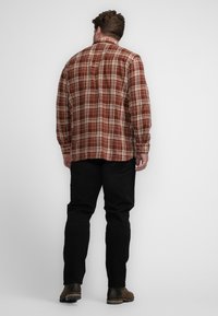 Rood en tan geruit flanellen overhemd, lange mouwen, met een knoopkraag. Draagend met zwarte broek en bruine laarzen, gezien vanuit de rug.