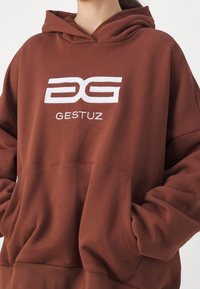 Osoba oblečená v hnědé mikině s kapucí, na hrudi s bílým logem "DG GESTUZ" a s rukama v přední kapse.