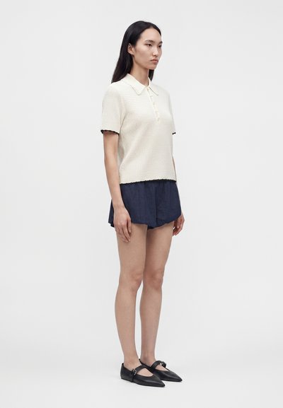 Femme debout portant un polo en maille blanc à manches courtes, un short bleu foncé et des ballerines pointues Mary Jane noires sur fond blanc.