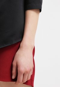 Top nero a maniche corte con finitura liscia, abbinato a una gonna rossa con texture. La mano è appoggiata sul tessuto della gonna.