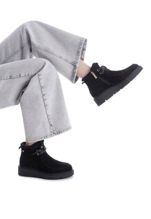 Botas de tobillo de ante negro con un cuello texturizado y esponjoso, que cuentan con un cierre de cordones y una suela de goma estriada. Llevadas con pantalones anchos grises.