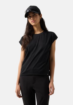 Jonge vrouw met lang donker haar, zwart petje, zwart shirt met korte mouwen en zwarte broek, staand tegen een eenvoudige lichte achtergrond.