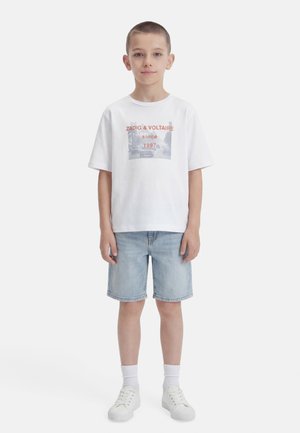 Junge steht, trägt ein weißes T-Shirt mit Grafik "ZADIG & VOLTAIRE seit 1997", helle Jeansshorts, weiße Socken und weiße Turnschuhe vor einfarbigem Hintergrund.