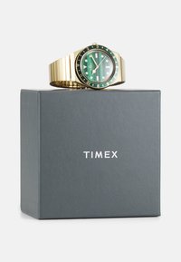 Montre en métal doré avec un cadran à rayures vertes, lunette noire et marqueurs blancs, présentée sur une boîte grise étiquetée "TIMEX".