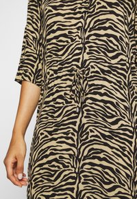 Abito color sabbia con motivo striato nero a zebra, maniche corte, dettaglio in vita con lacci e un tessuto leggermente texturizzato.