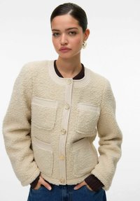 Krémová boucle bunda s texturovaným povrchem, s zlatými knoflíky, dvěma předními kapsami a kulatým výstřihem, ve spojení s hnědým topem.