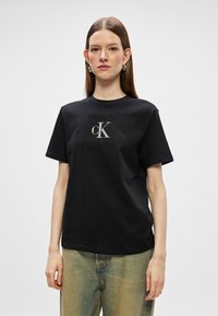 Crni pamučni majica kratkih rukava, s okruglim izrezom i srebrnim izvezenim "cK" logotipom na prednjoj strani. Model nosi široke svijetloplave traperice.