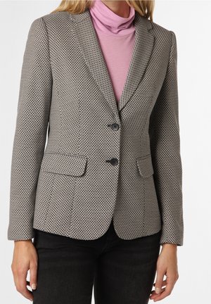 Mujer con blazer gris con estampado y botones negros sobre un jersey de cuello alto rosa y pantalones negros, mostrada desde los hombros hasta las caderas.