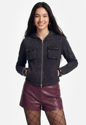 Ung kvinde med langt mørkt hår iført en sort hættetrøje med lynlås, piggede armbånd, burgunderfarvede lædershorts og sorte strømpebukser med diamantmønster, smilende.