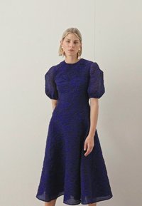 Um vestido de comprimento até ao joelho, com mangas bufantes, em tecido de renda azul profundo com um padrão floral texturizado e uma cintura ajustada.