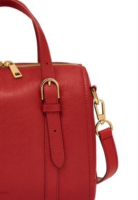 Un sac à main en cuir rouge présente une texture grainée, du matériel en ton doré, une fermeture éclaire sur le dessus et des poignées doubles avec une bandoulière ajustable.