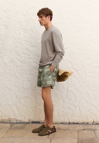 Hellgrauer Pullover, grüne gemusterte Shorts mit geometrischem Design, braune Sandalen mit Riemen und ein Strohhut, der in die Shorts gesteckt ist.