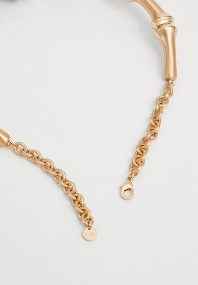 Gold metal chain with smooth cylindrical accents, featuring a lobster clasp and a small round tag. The overall design is sleek and modern.