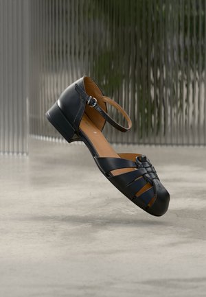 Sandale fermée en cuir noir avec sangle à la cheville et petit talon, présentée sur un fond flou à texture verticale et un sol en béton.