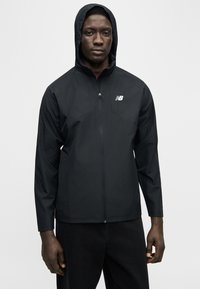 Άνδρας που φοράει μαύρο φούτερ New Balance με κουκούλα και μπροστινό φερμουάρ, στέκεται μπροστά σε απλό ανοιχτό φόντο.