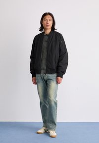 Giacca bomber nera con polsini a coste, tasca laterale con zip, indossata sopra un maglione grigio testurizzato e jeans azzurri chiari, abbinata a sneaker bianche.