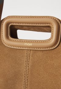 Brun ruskinds håndtaske med rektangulært læderhåndtag, detaljeret syning og lille guld "maje" logo under håndtaget.