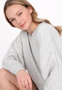 Šedá mikina s výstřihem do V a oversized rukávy, která má jemnou texturu. Modelka sedí a částečně ukazuje černé šortky.
