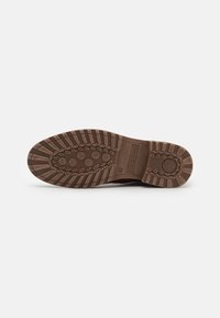 Semelle d'une chaussure marron avec des crampons profonds, des motifs circulaires antidérapants, et le nom de marque "bugatti" embossé près de la voûte plantaire.