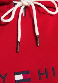 Hoodie rouge en tissu coton, avec une poche frontale, des cordons blancs avec embouts métalliques, et un détail de logo brodé dans des couleurs contrastées.