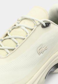 Αθλητικό παπούτσι σε κρεμ χρώμα με γλωττίδα από δίχτυ, κορδόνια ανοιχτού γκρι, λογότυπο κροκόδειλου στην πλευρά και μεταλλικό οφθαλμό κοντά στην κορυφή της τρύπας για τα κορδόνια.