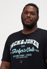 Melnas kokvilnas T-krekls ar baltu un gaiši zilu teksta dizainu: "JACK&JONES Surfer's Club 1990" un viļņu akcentiem un maziem simboliem.