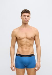 Niebieskie bokserki z czarnym pasem w talii z białym napisem marki "BOSS". Gładka faktura i dopasowany krój. Model stoi zwrócony przodem.