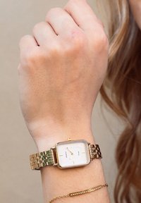 Montre en métal doré avec un cadran rectangulaire blanc, arborant des index horaires et un bracelet texturé, portée sur un poignet avec un délicat bracelet en or.