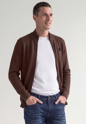 Mann trägt eine braune, strukturierte Reißverschlussjacke über einem weißen T-Shirt und blauen Jeans, steht mit den Händen in den Taschen, schaut zur Seite und lächelt.