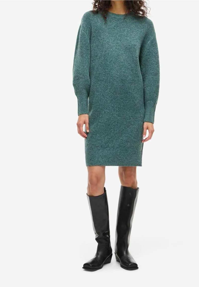 Femme portant une robe en maille à manches longues couleur sarcelle, associée à des bottes en cuir noires montant jusqu'aux genoux, debout devant un fond blanc.