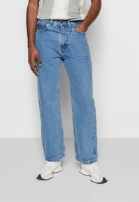 Homme portant un jean droit bleu clair, des baskets blanches et une chemise à manches courtes boutonnée, debout avec une main dans la poche.