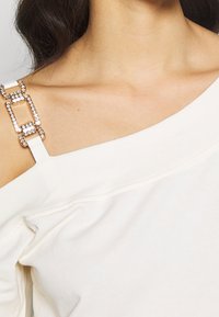 Vit av-axel topp med en slät textur och en dekorativ rem med rektangulära rhinestones.