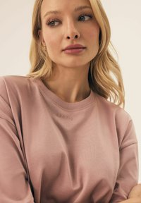 Jasnoróżowa, krótko rękawowa bluza z miękkiego materiału, z prążkowanym dekoltem i subtelnym tłoczeniem logo na klatce piersiowej.