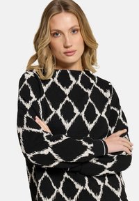 Vrouw met blond haar en blauwe ogen, gekleed in een zwarte trui met een wit ruitpatroon, met de armen gekruist voor een effen achtergrond.